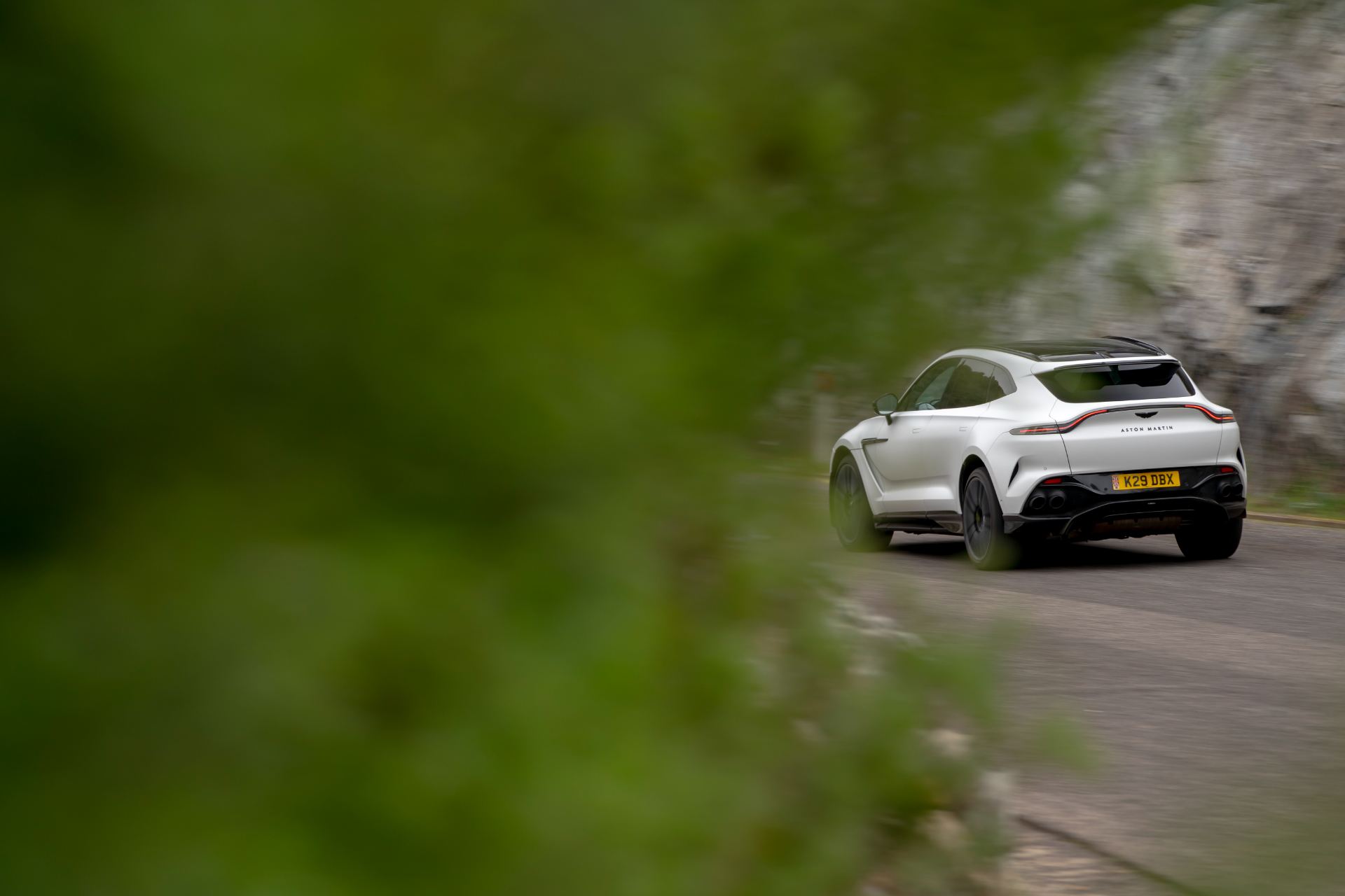 2026 aston martin dbx707 driving rear three quarter through foliage white