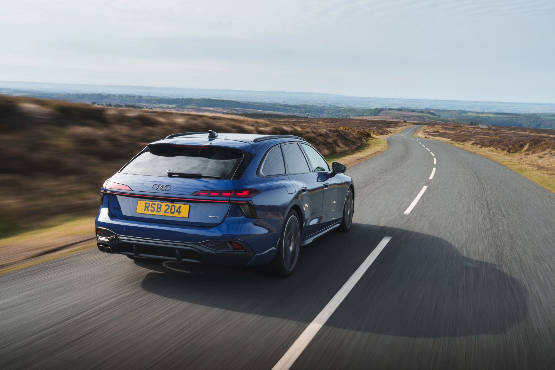 2026 audi a6 avant driving rear three quarter moorland road blue
