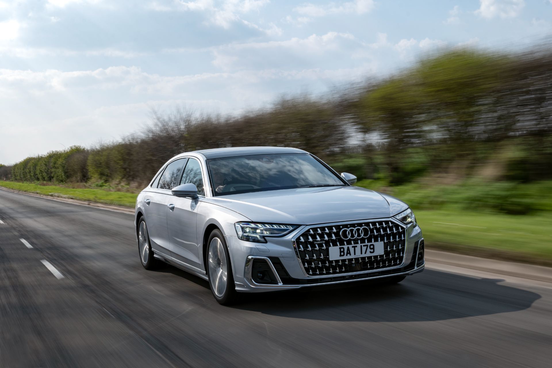 2025 audi a8 60 tfsi e driving front three quarter hedgerow silver